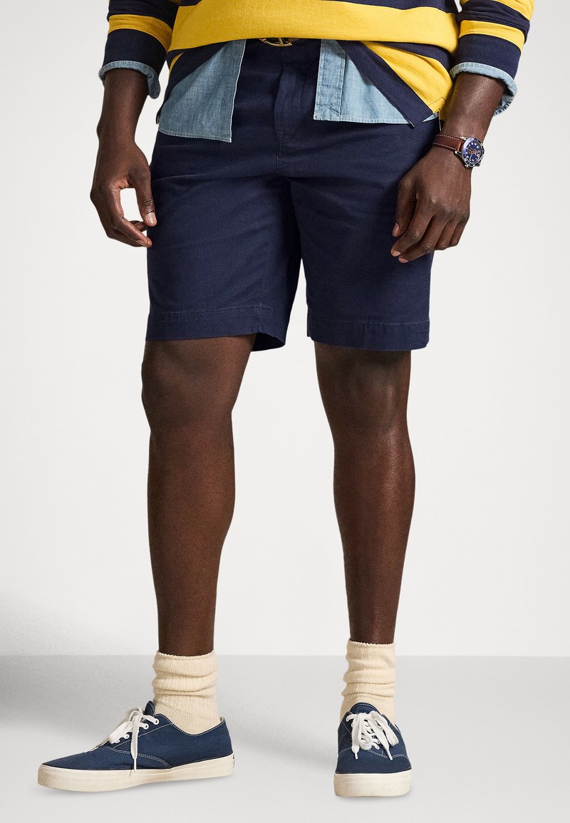 Tengerészkék pamut rövidnadrág, laza szabással, tengerészkék sportcipővel és krémszínű bokazoknival, amelyet színes csíkos hosszú ujjú inggel viselnek.