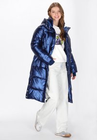 Hosszú, sötétkék puffermellény fényes felülettel, kapucnival és rugalmas mandzsettákkal. Egy fehér sweatshirt és világos farmernadrág fölé viselve, ezüst tornacipővel.