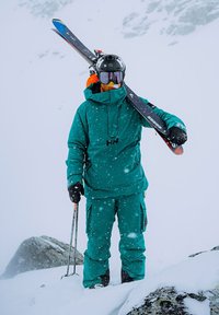 Een persoon in een groene skijack en -broek, met ski's in de hand, een helm en goggles, omringd door vallende sneeuw en rotsachtig terrein.