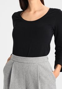 Schwarzes Langarmtop mit Rundhalsausschnitt, kombiniert mit hoch taillierten grauen plissierten Hosen, die seitliche Taschen und eine glatte Stoffoberfläche aufweisen.