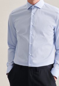 Camicia azzurro chiaro a maniche lunghe con bottone, dalla texture liscia, colletto a punta e bottoni bianchi, abbinata a pantaloni scuri.