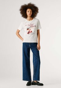 Maglietta bianca in cotone con grafica stampata con la scritta "PLAY THAT SOUND"; abbinata a jeans blu a gamba larga e mocassini neri.
