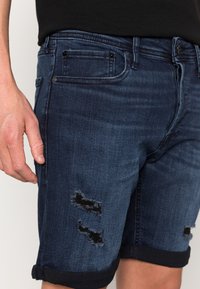 Tummansiniset farkkushortsit, joissa on kulutettuja kohtia, käännetyt lahkeet ja viisi taskua. Sileä rakenne, jossa on hieno haalistuma ja kontrastiväriset ompeleet.