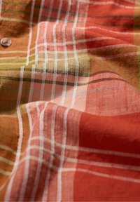 Gewebter Stoff in warmen Farben wie Rot, Orange und Grün, mit auffälligen weißen und schwarzen Karomustern sowie einem dekorativen Knopfdetail.