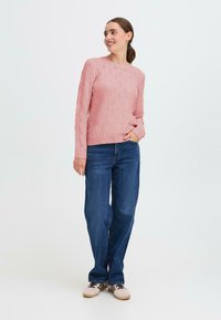 Maglione rosa a maglia con un motivo testurizzato, scollo rotondo e maniche lunghe, abbinato a jeans in denim blu dall'aspetto morbido e sneakers chiare.