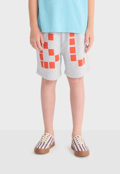 Enfant portant un short gris avec des motifs orange en blocs, un t-shirt bleu et des chaussures en toile rayées, se tenant contre un fond uni.