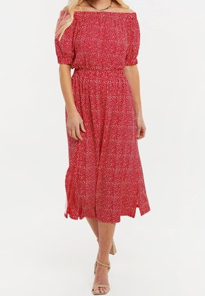 Woman wearing a red off-shoulder midi dress with white speckles, puff sleeves, elastic waist, and beige heeled sandals, standing on white background.