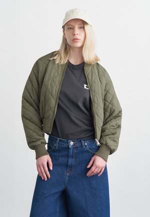 Jonge vrouw met blond haar, beige pet, gewatteerd olijfgroen jack, zwart T-shirt en wijde blauwe spijkerbroek, staand tegen een effen achtergrond.