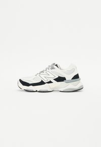 Sport shoes with a mix of white mesh and black suede. Thick sole, rounded shape, and gray laces. Features a prominent logo on the side.