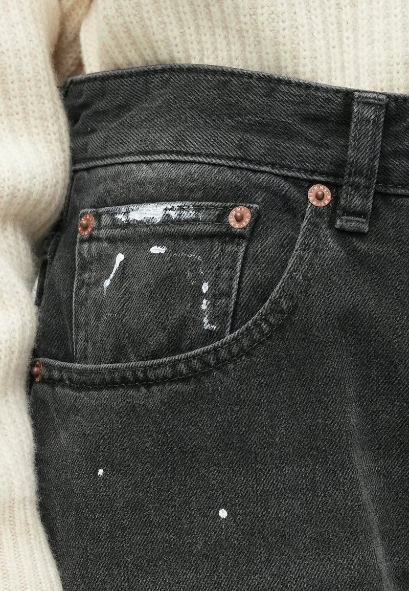 Donkergrijze denim jeans met een voorzak, voorzien van koperen klinknagels. Verfspetters op de stof. Zachte textuur met een iets versleten uiterlijk.