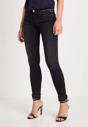 Jeans skinny negros con cintura alta, tejido de mezclilla elástico, desvanecimiento sutil, bolsillos frontales y dobladillos enrollados, combinados con sandalias de tacón negras.