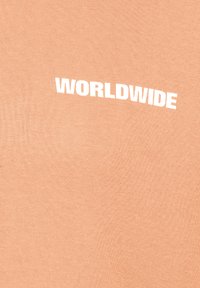 Pfirsichfarbiges Kurzarmshirt aus weicher Baumwolle. Das Wort "WORLDWIDE" ist in fettem Weiß auf der Brust aufgedruckt und wölbt sich über die Brust.