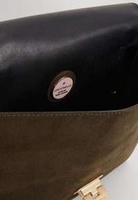 Zbliżenie na otwartą brązową torebkę ze skóry zamszowej i czarnej skóry z złotym zapięciem oraz okrągłą włoską metką marki Coccinelle wewnątrz.