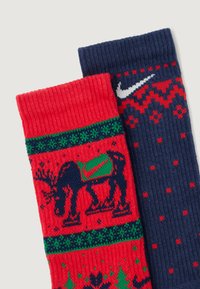 Adītas zeķes ar sarkanu dizainu, kurā ir brieža un Ziemassvētku raksti vienā pusē, un tumši zila puse ar ģeometriskiem akcentiem un Nike logo.