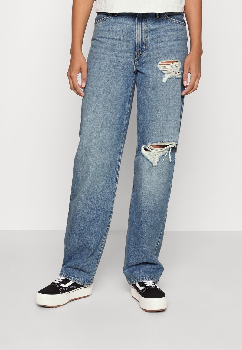 Personne portant un jean déchiré bleu clair et des baskets noir et blanc, debout devant un fond clair uni.