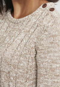 Sweter dzianinowy w beżowo-kremowej kolorystyce, z wzorem warkoczowym i dwoma brązowymi guzikami na ramieniu.