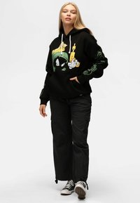 Felpa nera con design grafico che presenta un personaggio dei cartoni animati. Maniche lunghe con dettagli colorati. Abbinata a pantaloni cargo neri e sneakers.