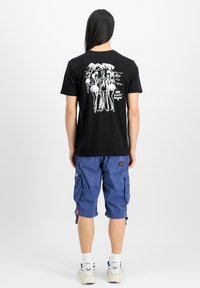 Melna kokvilnas T krekls ar baltu grafisko izdruku uz muguras, apvienots ar ziliem kravas šortiem un sporta apaviem.