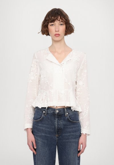 Weiße Bluse mit floralen Stickereien, langen Ärmeln und einem V-Ausschnitt, mit einem Peplum-Saum und durchsichtigen Stoff, kombiniert mit blauen Jeans.