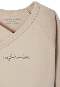 Beige Baumwoll-Babykleidung mit einem V-Ausschnitt. Mit blauer gestickter Schrift "100% fait maison". Glatte Textur und genähte Kanten.