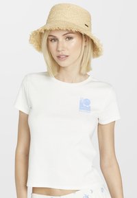 Witte cropped t-shirt met korte mouwen en een blauw graphic ontwerp op de linker borst. Draagt een natuurlijke rieten emmerhoed.