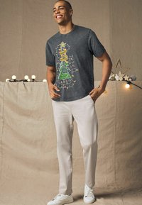 Sort t-shirt med et farverigt juletretræ design med lys. Bæres med lysfarvede bukser og hvide sneakers. Baggrunden har blødt lys.