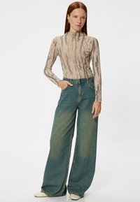 Langsleeve, beige top met golvende grijze patronen gecombineerd met wijdlopende, vervaagde teal jeans. Model poseert met één hand in de zak, minimalistische stijl.
