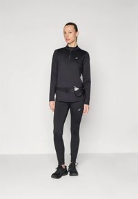 Donna in abbigliamento sportivo nero, tra cui una maglietta a maniche lunghe e leggings, con una cintura in vita che tiene una bottiglia d'acqua piccola, in piedi contro uno sfondo semplice.