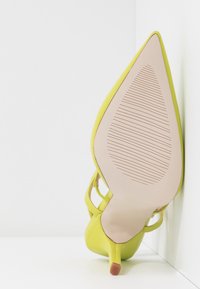 Chaussure à talon haut à bout pointu présentant un dessus en synthétique jaune vif, une semelle en caoutchouc nude avec des stries et de fines sangles à la cheville.