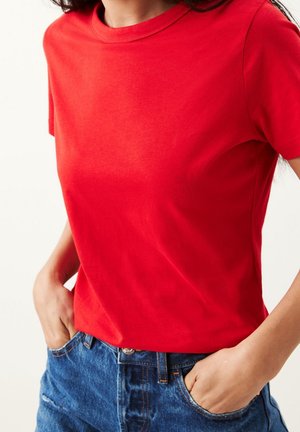 Mujer con camiseta roja de manga corta lisa metida dentro de unos vaqueros azules, con las manos en los bolsillos, frente a un fondo blanco.