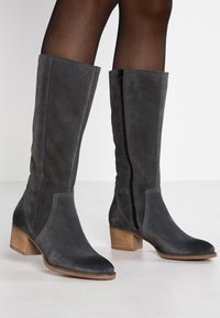 Szare zamszowe botki za kolano z ostro zakończonym noskiem i drewnianym obcasem w kształcie bloku. Posiadają boczny suwak i szczegóły w kontrastowym sticu.