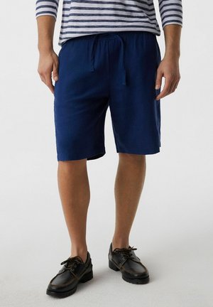 Mężczyzna w granatowych szortach ze ściągaczem, czarnych skórzanych butach typu boat shoes i w paskowanej koszuli z długim rękawem, stoi na jasnej podłodze.