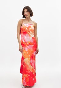 Moteris vilki be rankovių ilgą suknelę su oranžiniais ir rožiniais gėlių raštais, stovinti prieš paprastą baltą foną.