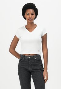 White cropped ribbed t-shirt with a V-neckline and short sleeves, paired with high-waisted dark grey jeans. Subtle logo tag at the hem.