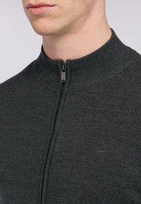 Pánský tmavě šedý pletený svetr s vysokým límcem, zipem vpředu a jemným logem. Textura je jemná a žebrovaná.