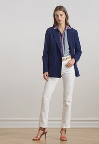 Marineblå pinstripet blazer over en stripete skjorte, hvite slim-fit bukser og brune hæl-sandaler med røde aksenter. Glatte teksturer og skreddersydd passform.