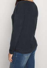 Dark navy knitted sweater with a textured finish, featuring long sleeves and a curved hem. Paired with blue denim jeans.
