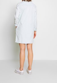 Light blue denim shirt dress with long sleeves, pockets, and a button-down design. Ribbed texture, knee-length, paired with white sneakers.