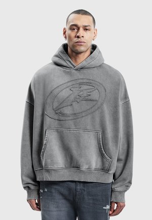 Hombre con el pelo corto y pendientes, vestido con una sudadera con capucha gris descolorida con un bolsillo frontal y un logo oval bordado.