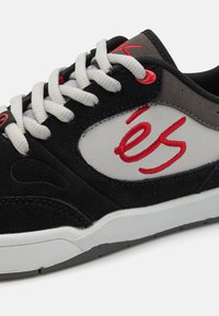 Baskets en suédine noire avec accents gris, logo rouge brodé, lacets blancs, bout rond et semelle texturée. Présente un intérieur rembourré.