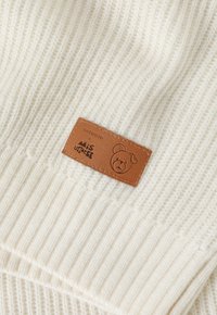 Pulover tricotat de culoare crem, cu o textură ribbed, având un eticheta din piele maro, embosată cu un grafic de urs și detalii ale mărcii.