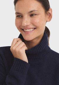 Marineblauer Rollkragenpullover aus weichem Material mit geripptem Design an Kragen und Ärmelbündchen; Modell hält sanft den Kragen.