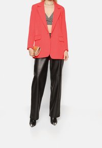Blazer rosso su misura con rever a stampa, due tasche frontali, abbinato a pantaloni a gamba larga in pelle sintetica nera e décolleté nere a punta.