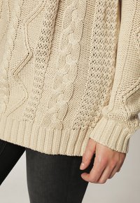 Beżowy sweter dzianinowy z mieszanką wzorów warkoczowych i otwartych, z ściągaczami na mankietach i u dołu. Tkanina wydaje się miękka i teksturowa.
