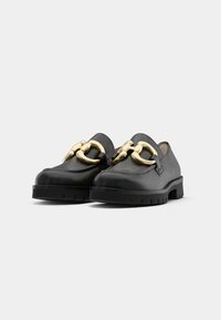 Zwarte leren loafers met chunky zolen en grote glanzende gouden kettingversieringen aan de voorkant, weergegeven op een witte achtergrond.