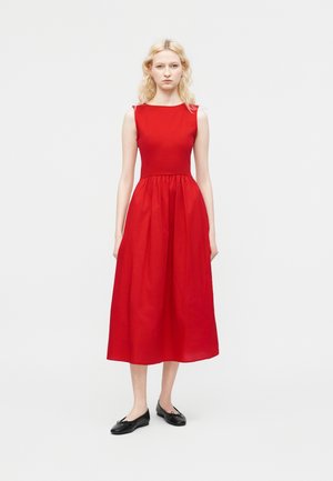 Ξανθιά γυναίκα όρθια, φοράει αμάνικο κόκκινο μεσαίο φόρεμα με εφαρμοστή κορυφή και μαύρα μπαλαρίνες, μπροστά σε απλό φωτεινό φόντο.