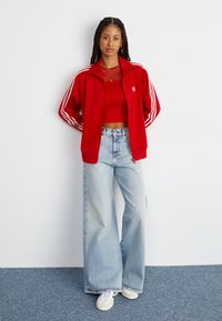 Giacca a tuta rossa con righe bianche, top corto rosso, jeans a gamba larga blu chiaro e sneakers bianche. Tessuto liscio e vestibilità rilassata.