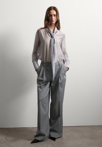 Blusa grigia chiara trasparente con colletto annodato, maniche lunghe e polsini arricciati abbinata a pantaloni grigi larghi e a vita alta e tacchi neri a punta.