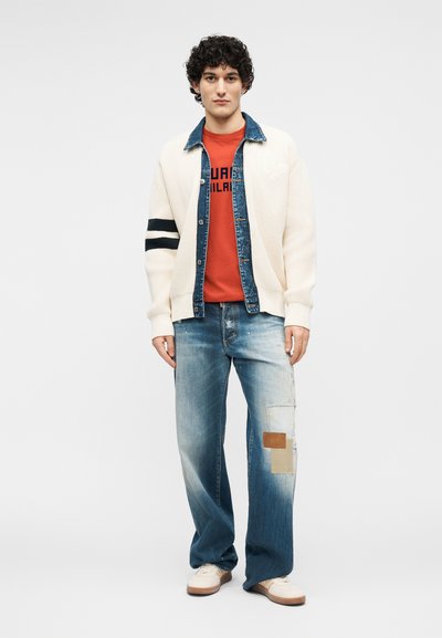 Jeune homme aux cheveux bouclés portant un cardigan blanc à rayures noires, un t-shirt rouge à motif, un jean bleu avec des patchs et des baskets blanches.