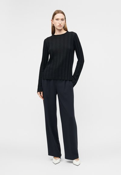 Femme debout, portant un haut noir à manches longues côtelé, un pantalon large bleu marine, des chaussures blanches pointues et des boucles d'oreilles dorées en forme d'anneau.